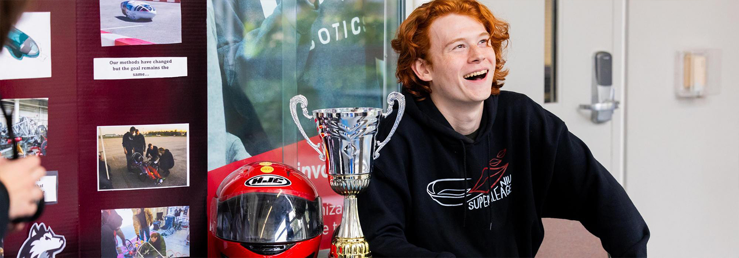 Student sitting beside a trophy and red racing helmet, smiling at a display about a vehicle competition project.